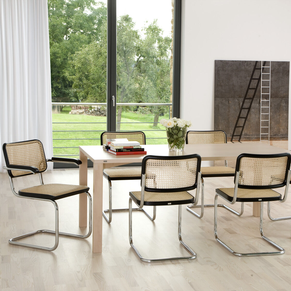 Ein Esszimmer mit modernen Thonet »S 64« Freischwingerstühlen aus Stahlrohr mit Rattan-Sitzfläche um einen rechteckigen Holztisch. Im Hintergrund ist eine große Fensterfront mit Blick auf einen grünen Garten sowie Kunst an der Wand sichtbar. Der Raum ist hell und minimalistisch eingerichtet.