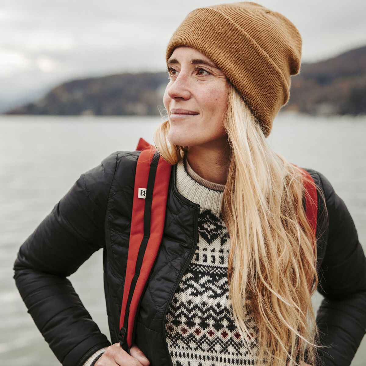 Eine Frau mit langen blonden Haaren trägt einen braunen Hut, einen schwarz-weißen Pullover mit skandinavischem Muster und einen schwarzen Outdoor-Jacke. Sie hat einen roten Rucksack »Wanaka« auf dem Rücken. Im Hintergrund ist ein See mit Bergen und nebelverhangener Himmel zu sehen.