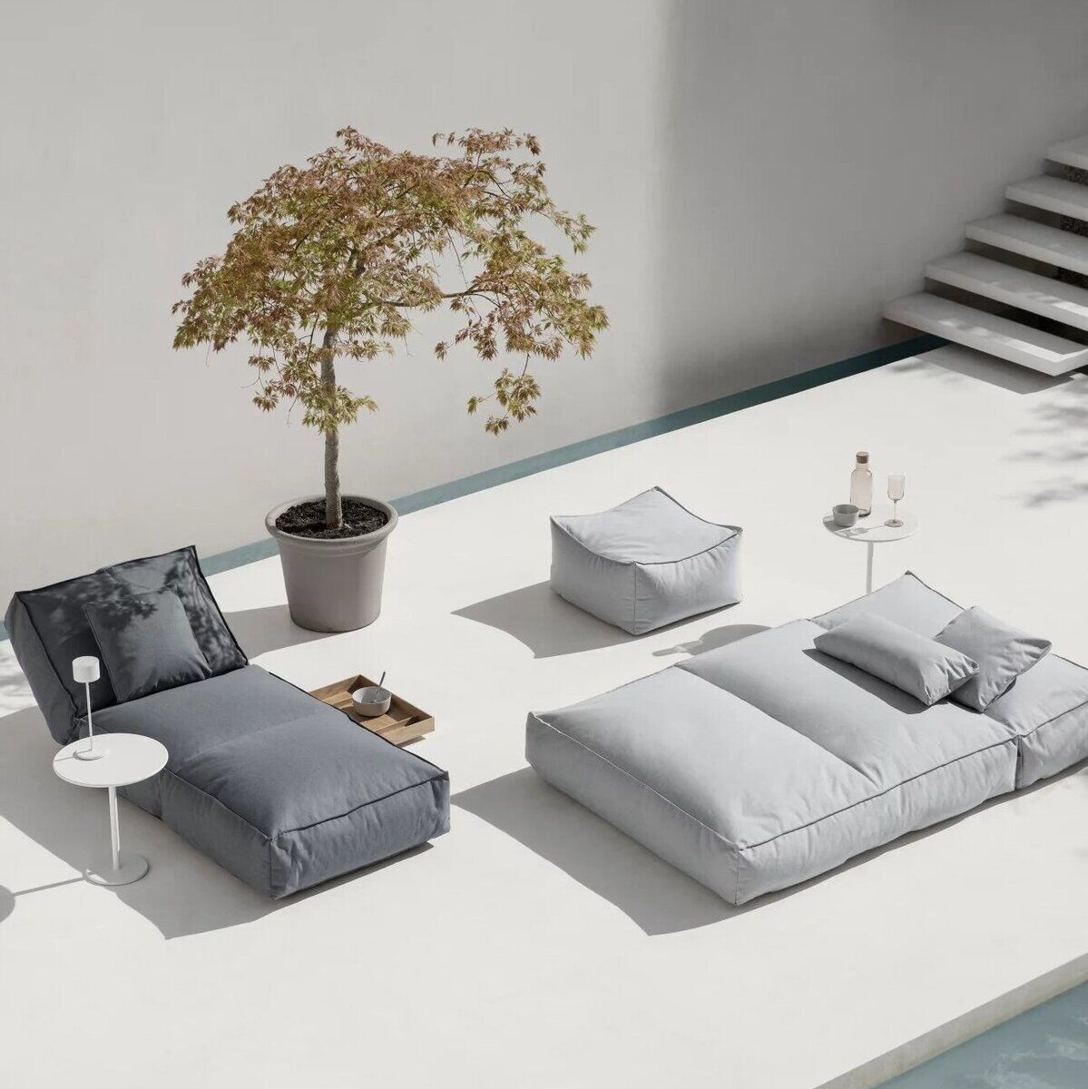 Ein minimalistisches Outdoor-Bett »STAY« in Grautönen, 80 x 190 cm, steht auf einer weißen Terrasse neben einem identischen grauen Stuhl und einer Pflanze in einem Blumentopf. Es gibt auch zwei runde Beistelltische mit Getränken und eine graue quadratische Sitzgelegenheit.