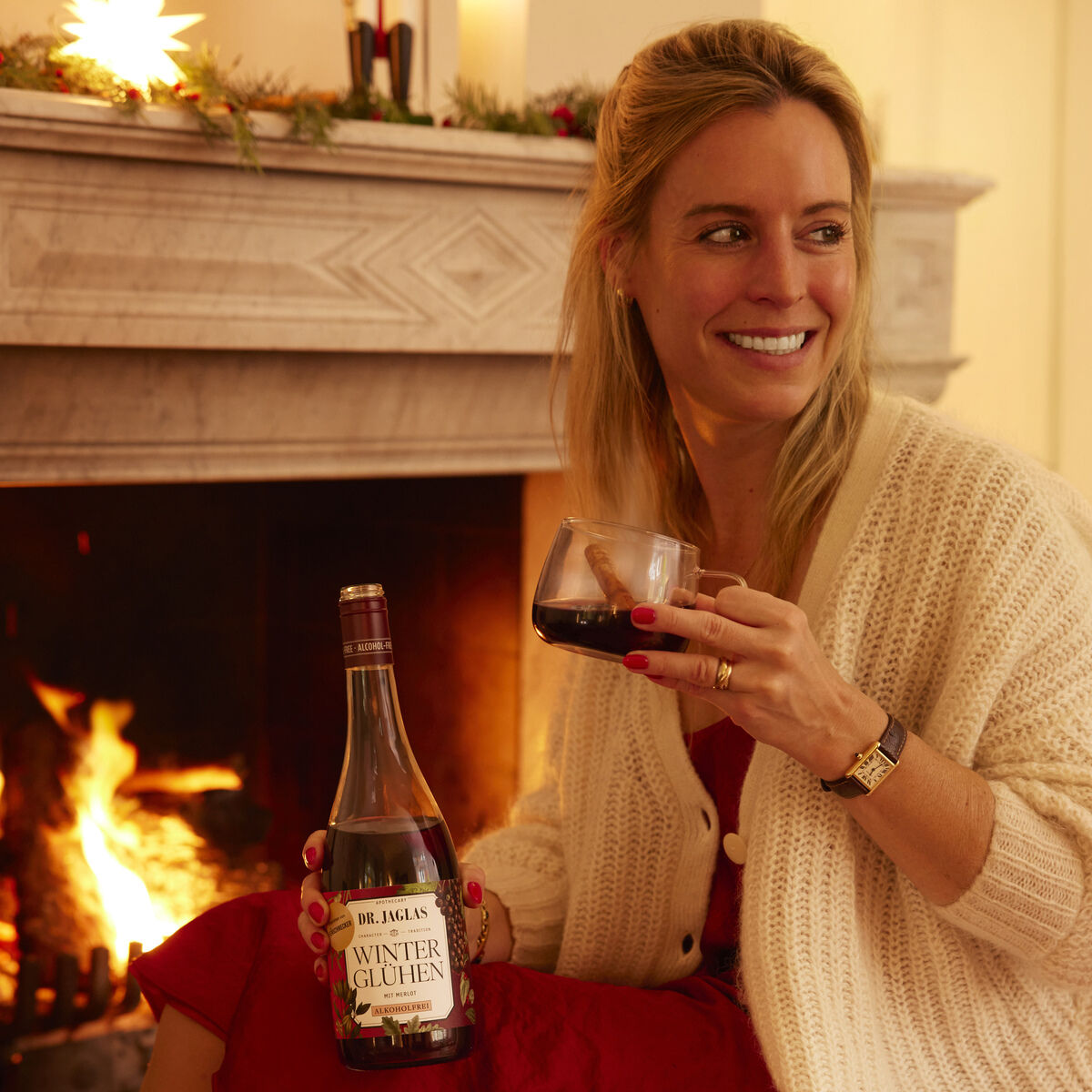 Frau sitzt gemütlich vor einem brennenden Kamin und hält ein Glas roten Winterglühen Merlot Wein, während sie lächelt. Im Hintergrund sind eine festliche Dekoration und Kerzen auf einem Kaminsims zu sehen. Die Szene vermittelt eine warme und festliche Winteratmosphäre.
