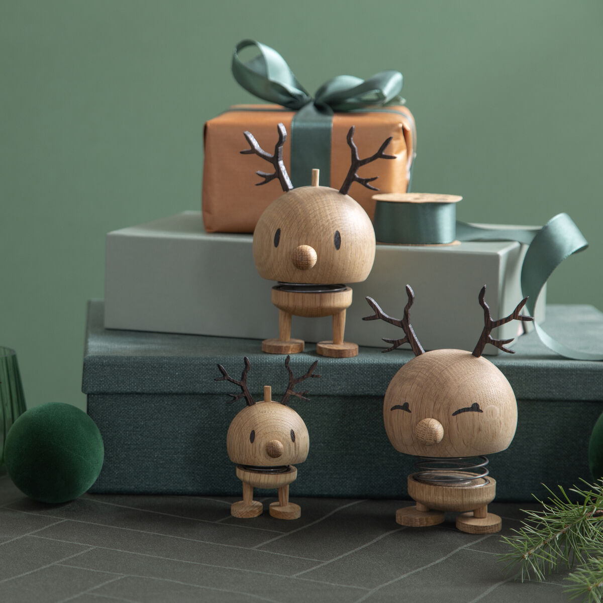 Vier Hoptimist Rentier Bumble Figuren aus Eichenholz mit schwarzen Geweihen stehen vor türkisgrünen Geschenkboxen und Tannenzweigen, auf einem grauen Tisch, mit einem festlichen und minimalistischen Hintergrund.