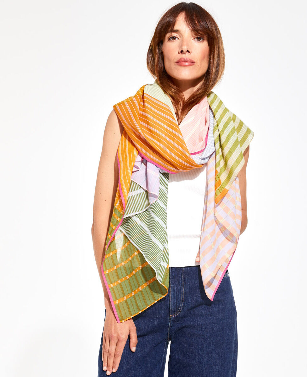 Frau trägt ein bunt gestreiftes Baumwolltuch mit verschiedenen Farben, darunter Orange, Grün, Pink und Gelb, drapiert über ein weißes Oberteil und blaue Jeans.