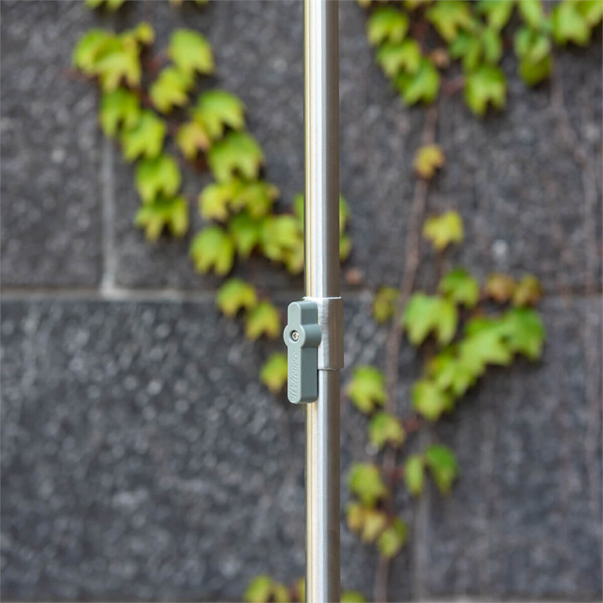 Eine Gartendusche »Serpentine« aus rostfreiem Stahl, abgebildet im Garten vor einer grauen Steinwand mit grünen Pflanzen im Hintergrund. Die Dusche verfügt über einen verstellbaren Griff.