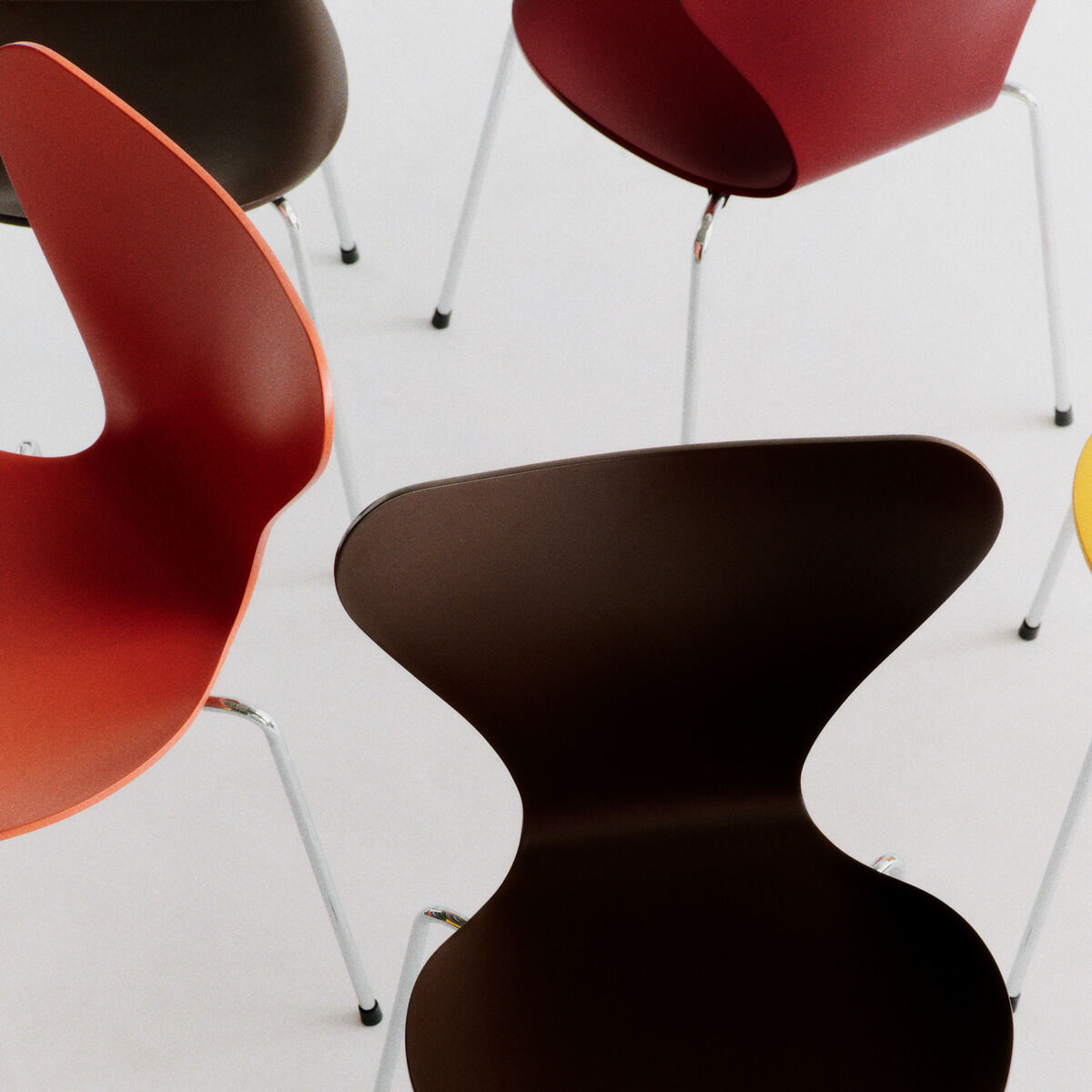 Mehrere farbige Stühle der Serie 7, Edition Verner Panton von Fritz Hansen sind locker angeordnet. Die Sitzschalen sind in Orange, Gelb, Dunkelbraun und Rot lackiert. Die Beine bestehen aus glänzendem Stahlrohr. Die Szene zeigt modernes Design auf hellem Hintergrund.