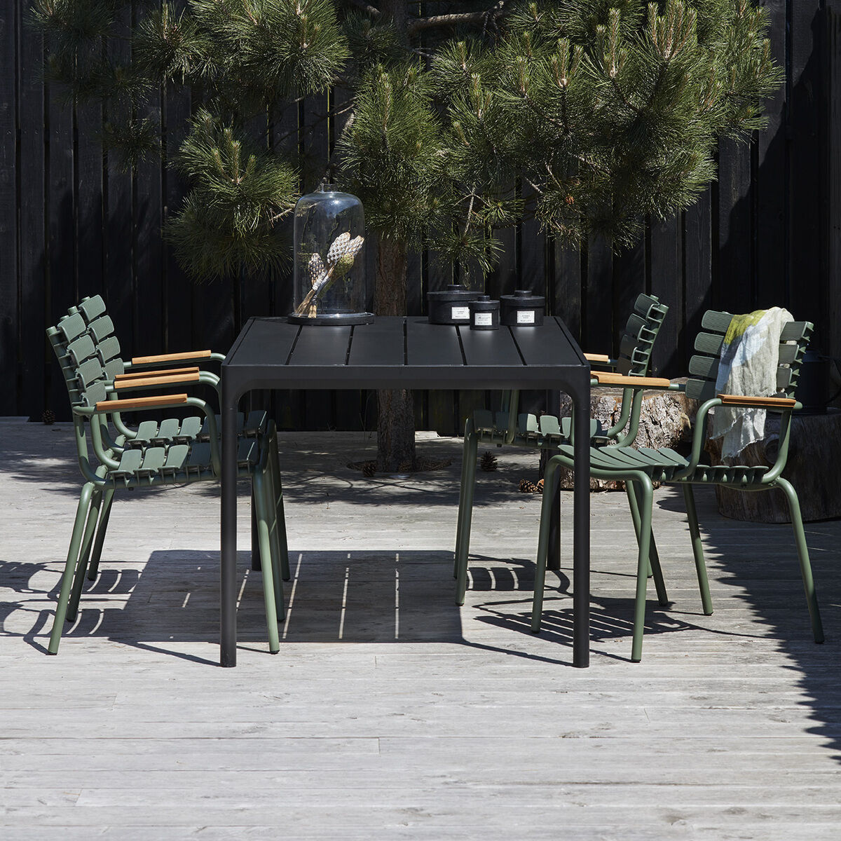 Draußen in einem Gartenbereich stehen vier grüne Stühle mit braunen Armlehnen um einen schwarzen Tisch, umgeben von einem hölzernen Zaun und einem Nadelbaum.