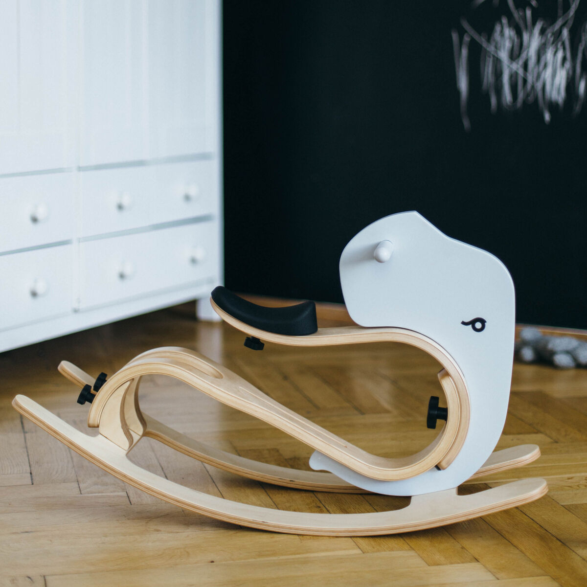 Ein kindgerechtes Schaukeltier in Elefantenform aus Holz mit hellen Naturtönen und weißen sowie schwarzen Akzenten steht auf einem Holzboden vor einer schwarzen Wand. Das Design ist modern und minimalistisch, ideal als Erweiterung für ein Balance Bike im Kinderzimmer.