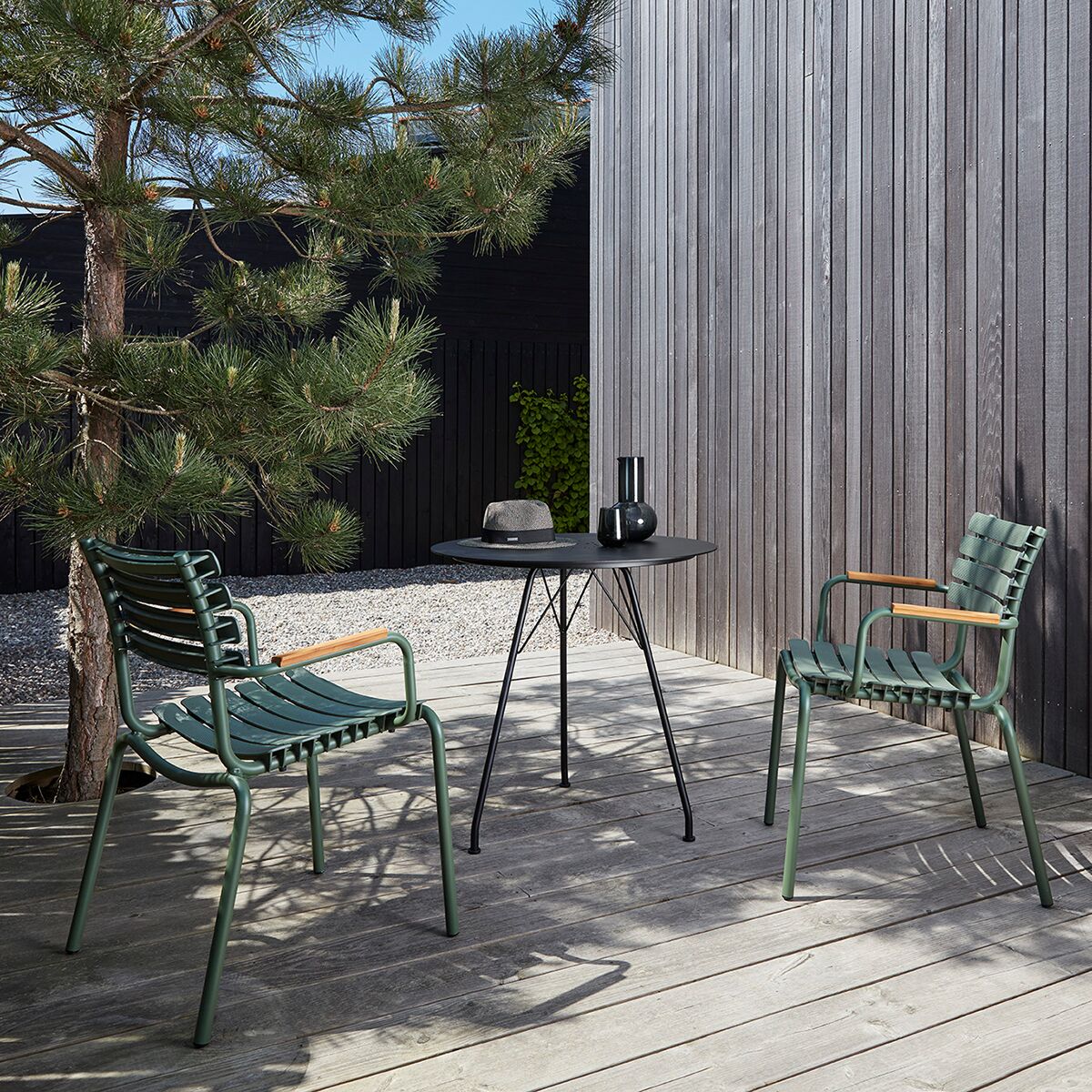 Auf einer modernen Holzterrasse stehen zwei olivgrüne Stühle der Serie »RECLIPS« von Houe mit breiten Kunststofflamellen und Armlehnen aus Bambus. Zwischen ihnen ein schlichter runder schwarzer Tisch, darauf eine schwarze Vase sowie eine Schale. Im Hintergrund steht eine Kiefer vor einer grauen Holzfassade.