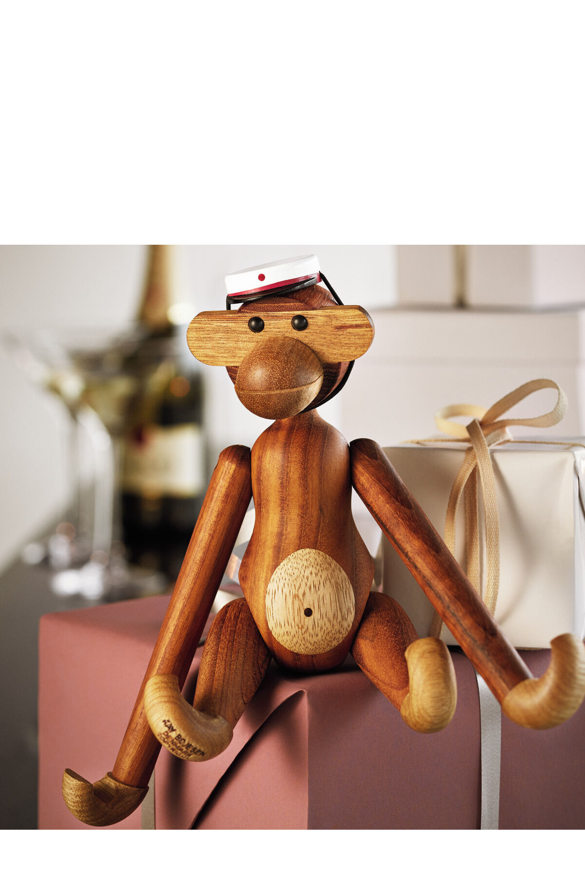 Die Holzfigur »Affe« von Kay Bojesen sitzt auf einem Geschenkstapel. Im Hintergrund sind weitere Geschenke, eine Champagnerflasche und Gläser zu sehen. Die Figur ist aus verschiedenfarbigem Holz gefertigt und trägt ein kleines Mützchen.