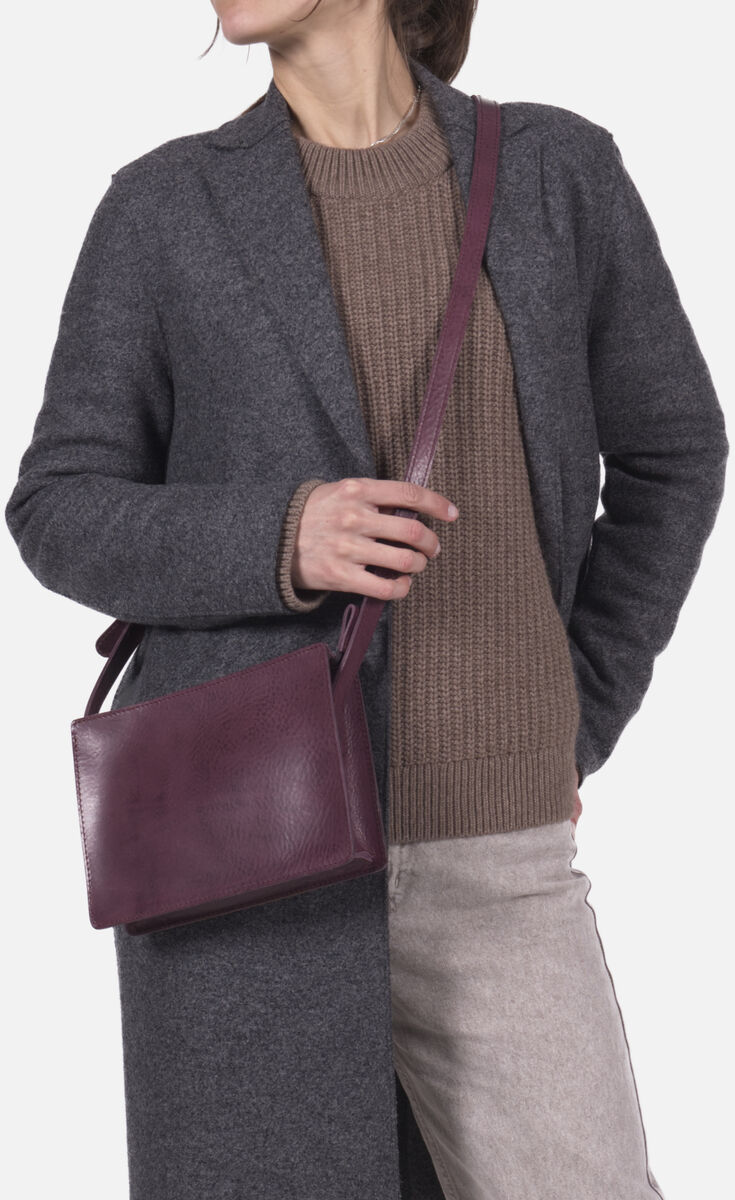 Frau trägt eine dunkelviolette Kernleder-Umhängetasche der Marke „Sas“ über der Schulter. Sie trägt einen langen grauen Mantel, einen braunen Strickpullover und beige Hose. Die Ledertasche hat ein schlichtes und elegantes Design. Hintergrund ist neutral weiß.