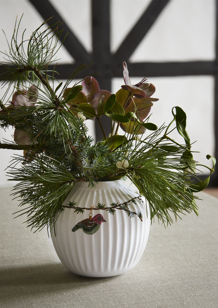 Weiße Hammershøi-Vase mit vertikalen Rillen, dekoriert mit einem grünen Nadelzweig und Blättern. Eine kleine Vogelillustration befindet sich auf der Vase. Hintergrund ist mit einem Holzmuster dekoriert. Perfekt für weihnachtliche oder winterliche Dekoration.