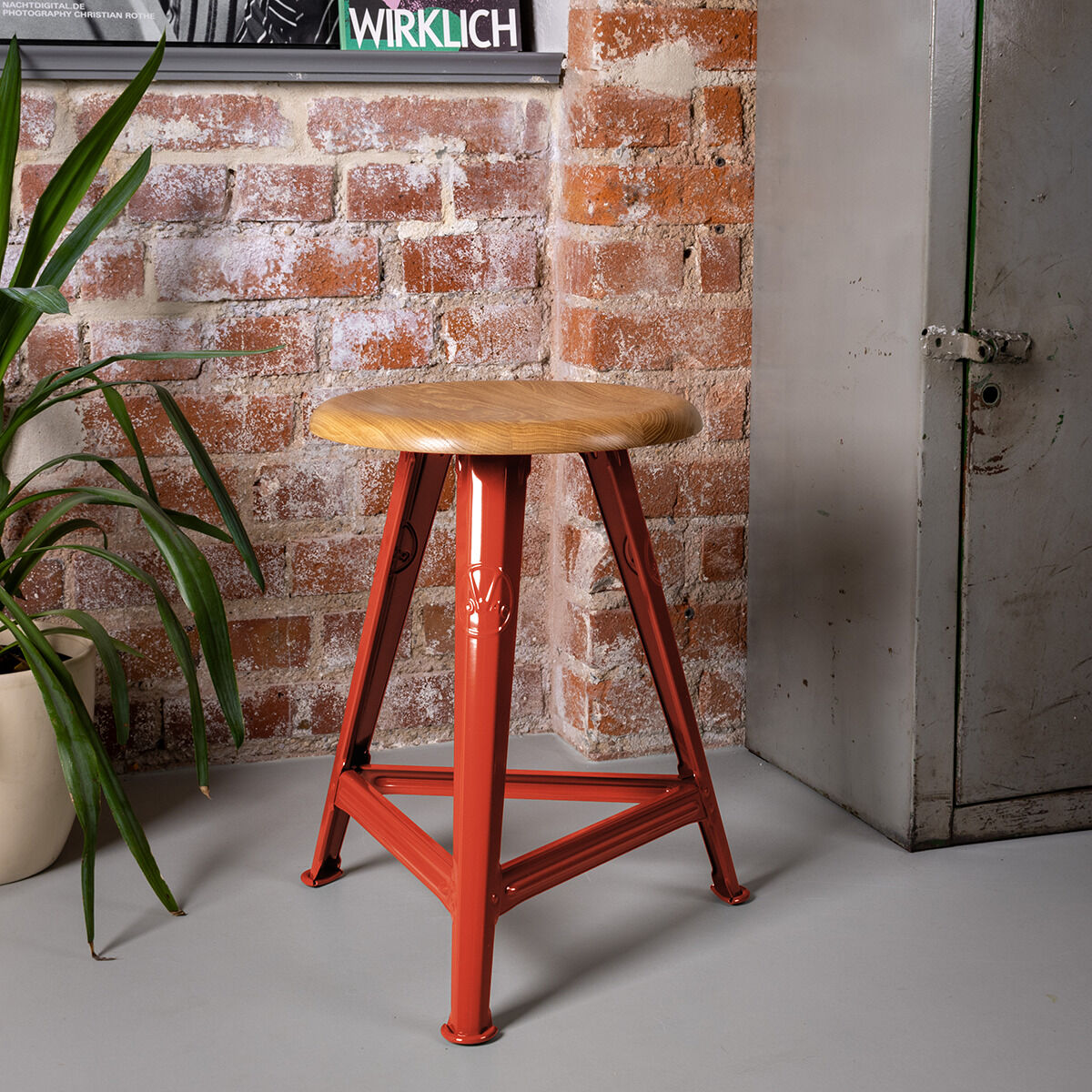 Hocker aus der ZEIT-Sonderedition Rowac-Schemel »Red Brick« mit rotem Metallgestell und einer Holzsitzfläche, stehend auf einem Boden aus grauem Beton, neben einer Pflanze in einem beigen Übertopf und vor einer Backsteinwand.