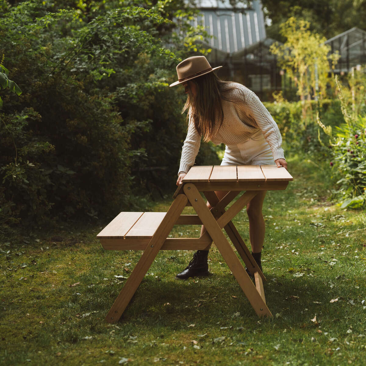 Im Bild steht eine Frau mit Hut im Garten neben einer klappbaren Tischbank namens »Folding Picnic Table« der Marke Weltevree. Die Tischbank ist aus braunem Bambus und steht auf einer grünen Rasenfläche, umgeben von üppigem Grün und Pflanzen im Hintergrund.
