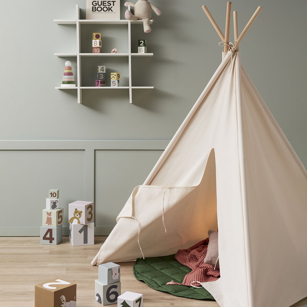 Ein 160 cm hohes cremeweißes Tipi-Zelt steht in einem modern eingerichteten Kinderzimmer mit Holzboden. Im Hintergrund befinden sich bunte Holzklötze mit Zahlen, Wandregale mit Dekoartikeln und ein Plüschhase. Das Zelt ist offen und mit Kissen sowie einer Decke gemütlich gestaltet. Das Foto vermittelt eine freundliche und wohnliche Atmosphäre für Kinder.
