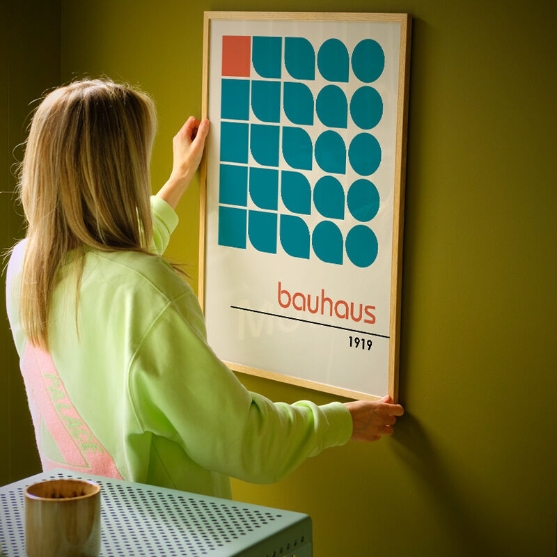 Eine Person hängt ein gerahmtes Poster mit geometrischen Formen an eine grünliche Wand. Das Kunstwerk ist im Bauhaus-Stil gehalten und beinhaltet Kreise und Quadrate in Blau und Rot mit der Aufschrift 'bauhaus 1919'.