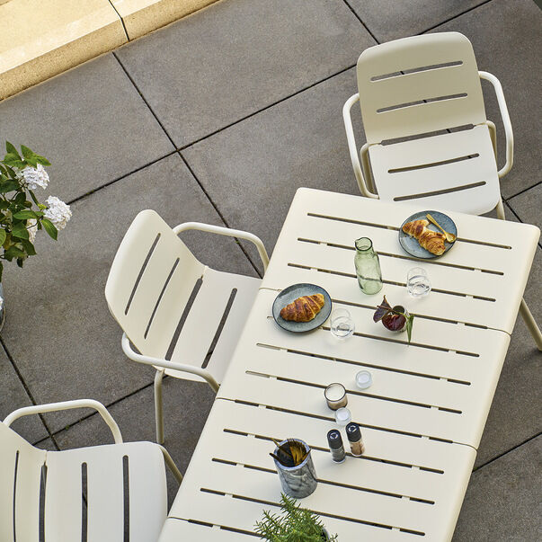 Ein Außenbild von mehreren weißen Aluminiumstühlen und Tischen, die auf einer Terrasse arrangiert sind. Auf den Tischen stehen Teller mit Croissants und Tassen mit Getränken. Im Hintergrund sind einige Pflanzenarrangements zu sehen.