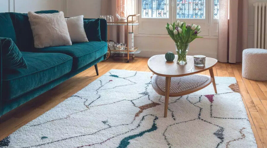 Wohnzimmer mit grünem Samtsofa, hellem Teppich mit abstraktem Muster, rundem Holztisch und Vase mit weißen Tulpen