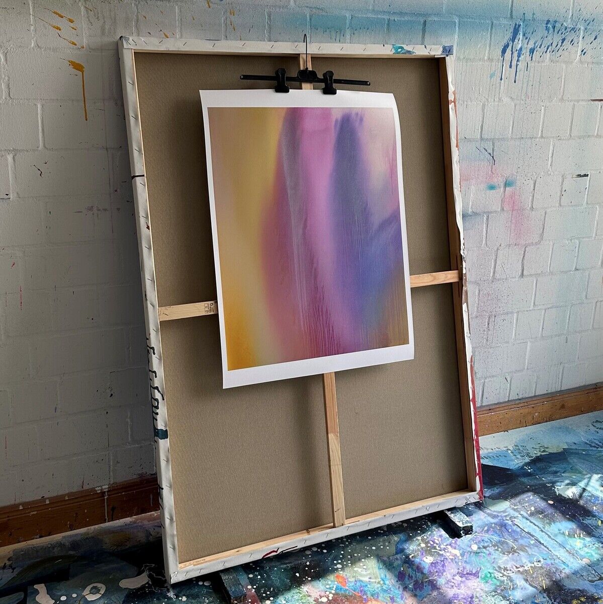 Ein abstraktes Kunstwerk der ZEIT Sonderedition von Paul Schrader mit dem Titel »Miami No. 3«. Das Bild zeigt weiche Übergänge von lila, rosa und gelben Farben auf einer Leinwand, die an einer Staffelei vor einer strukturierten weißen Wand steht. Der Boden ist farbenfroh mit Farbflecken bedeckt.