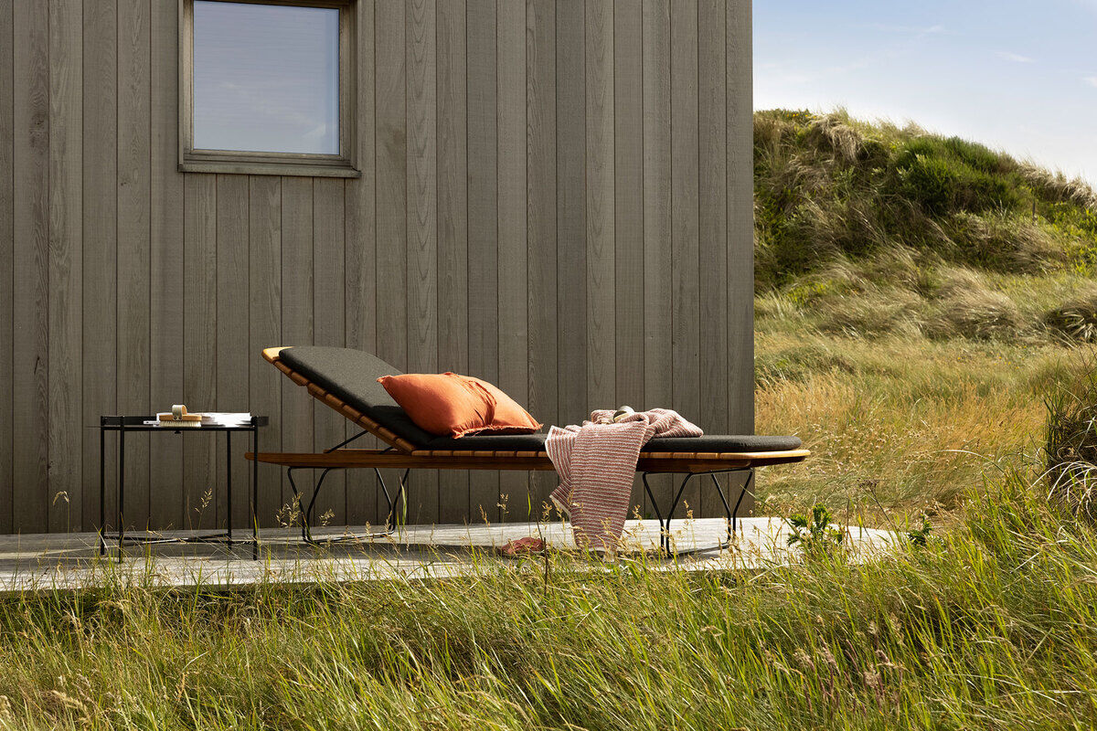 Eine stilvolle Sonnenliege mit schwarzer Auflage und orangefarbenem Kissen steht vor einer grauen Holzverkleidung im Garten/Outdoor-Bereich. Ein kleiner Beistelltisch mit Frühstück steht daneben. Im Hintergrund ist eine grüne Rasenfläche zu sehen.