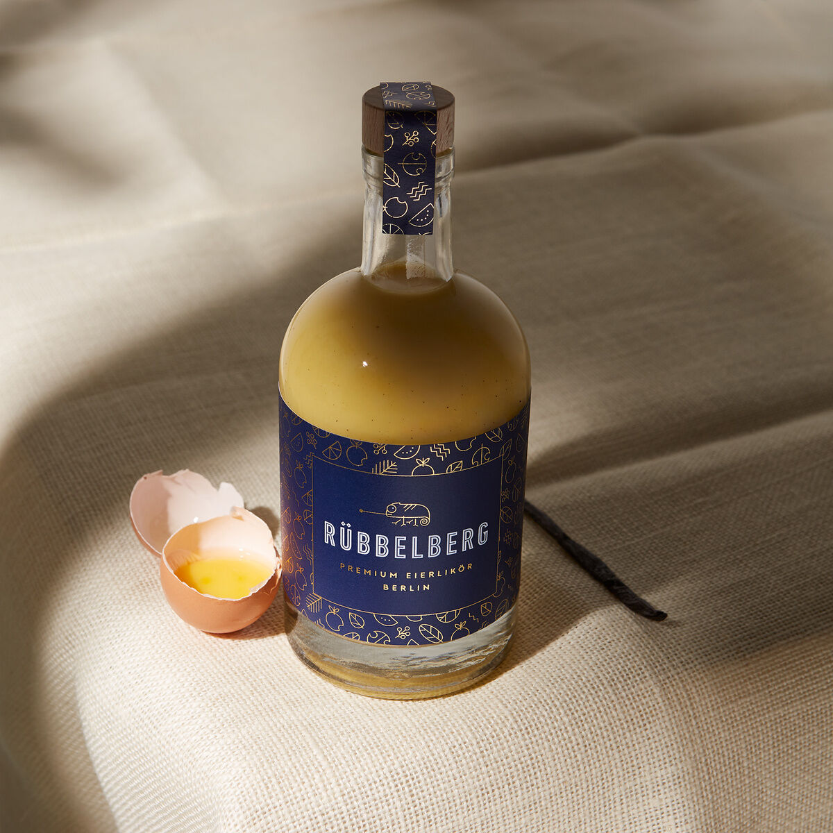 Eine bauchige Glasflasche »Premium Eierlikör« von Rübbelberg mit dunkelblauem Etikett und Korkverschluss steht auf beige strukturiertem Stoff. Daneben liegt eine aufgeschlagene Eierschale und eine Vanilleschote, beide werden von Sonnenlicht akzentuiert.