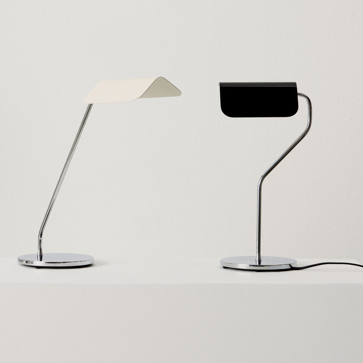 Zwei moderne Tischlampen mit minimalistischem Design auf einem weißen Tisch. Eine Lampe ist weiß, die andere schwarz, beide haben einen Chromfuß und elegante, schlanke Linien.