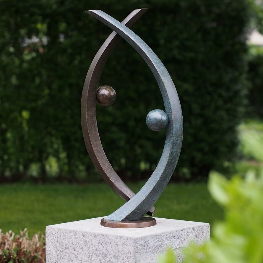 Abstrakte Gartenskulptur mit zwei gebogenen, sich kreuzenden Figuren aus Metall, die auf einem Steinsockel stehen. Die Skulptur steht in einem grünen Garten mit Büschen und Bäumen im Hintergrund.