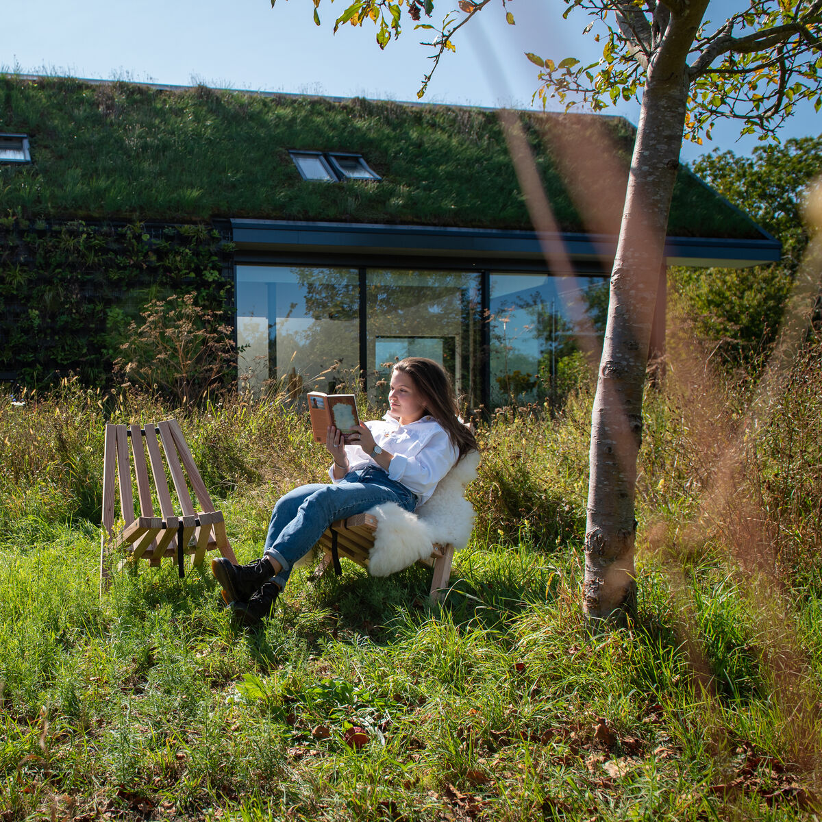 Eine Person sitzt entspannt auf einem mit Schafsfell bezogenen Stuhl im Garten vor einem modernen Haus mit begrüntem Dach, umgeben von üppiger Vegetation und Sonnenlicht.