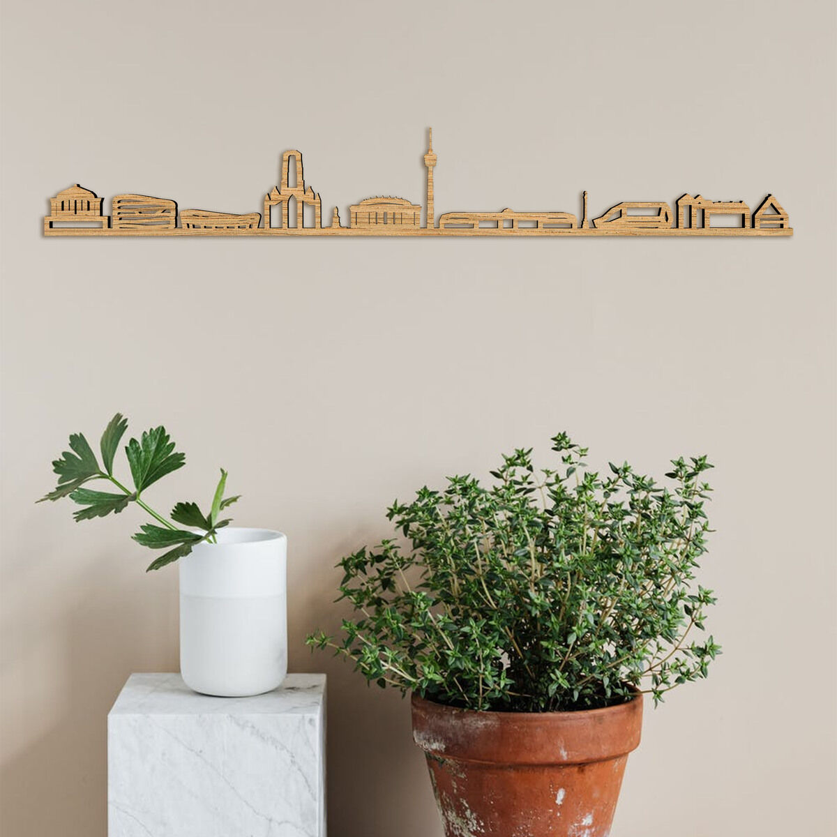 Holzkunstwerk der Skyline von Stuttgart, an einer beigen Wand aufgehängt, mit einem weißen Sockel und einem Blumentopf mit grünen Pflanzen darunter.