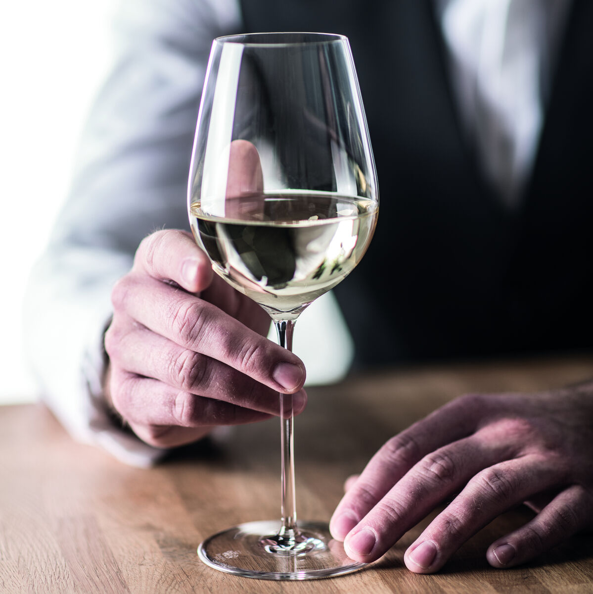 Ein Mann in einem schwarzen Anzug hält ein Glas Weißwein vorsichtig in der Hand. Das Glas steht auf einer Holzoberfläche.