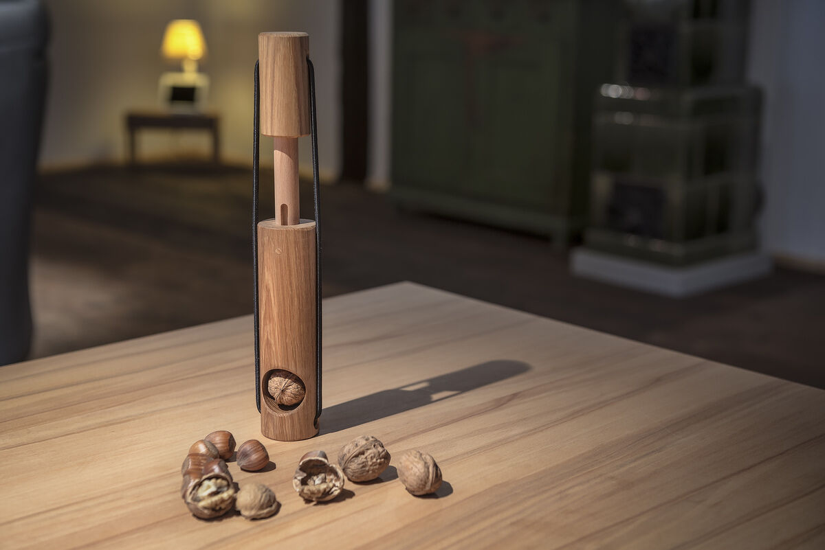 Ein eleganter Nussknacker aus Holz mit einem ergonomischen Design steht auf einem Holztisch. Um den Nussknacker herum liegen geöffnete und ganze Walnüsse. Im Hintergrund sind unscharfe Details eines Raumes mit einem Sofa und einer Lampe sichtbar.