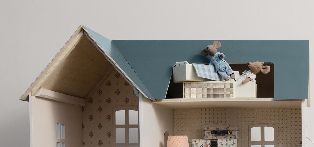 Ein Puppenhaus mit zwei Stoffmäusen auf einem Hochbett. Das Puppenhaus ist detailliert gestaltet mit einem Bett mit karierter Bettwäsche und weiteren Möbelstücken. Die Wände des Puppenhauses sind hell und das Dach ist dunkelblau. Der Raum sieht gemütlich und einladend aus.