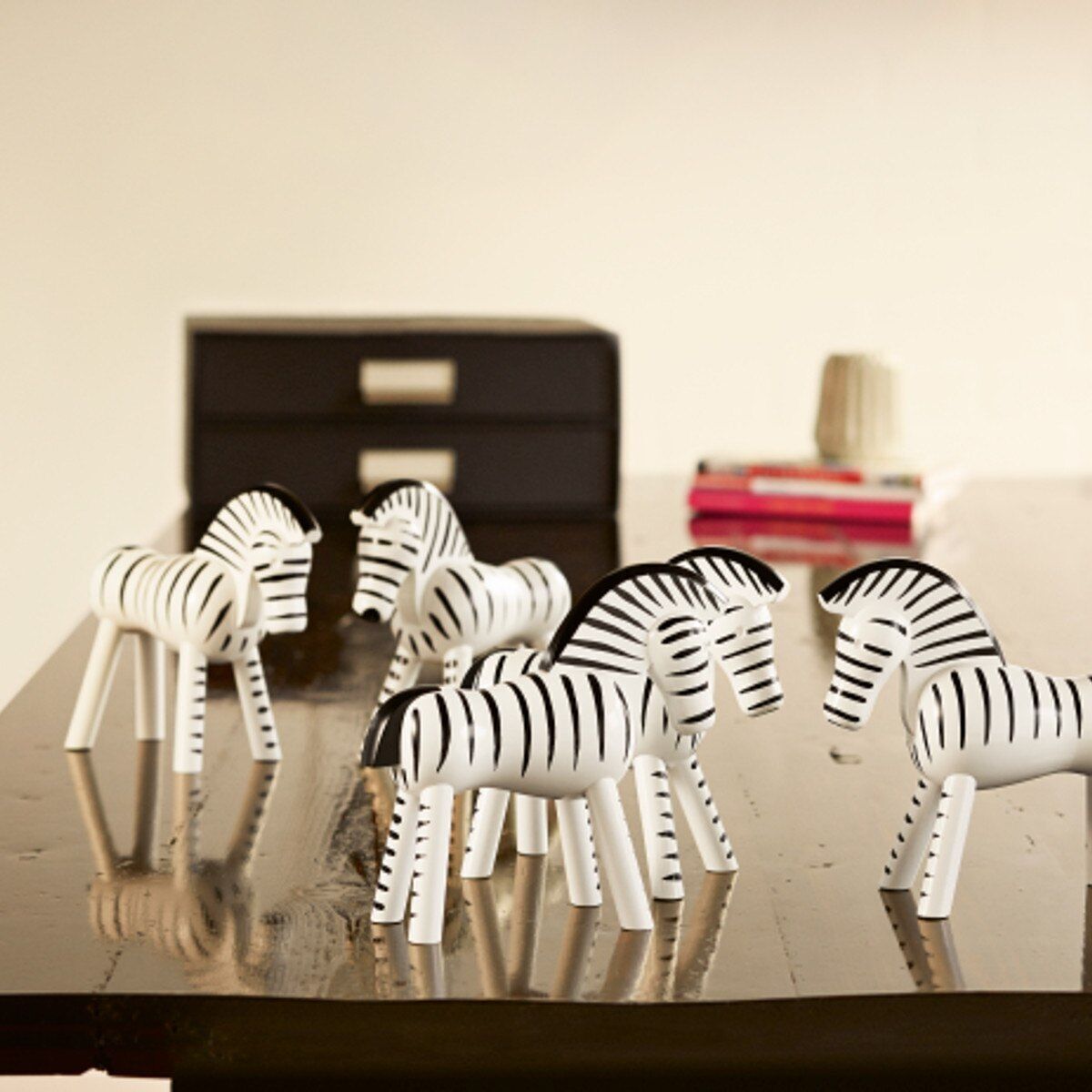 Mehrere Holzfiguren in Form von Zebras stehen auf einem Tisch. Die Figuren sind in Schwarz-Weiß gehalten und sind ein Kunsthandwerk von Kay Bojesen. Im Hintergrund sind unscharfe Gegenstände und eine Wand zu sehen.