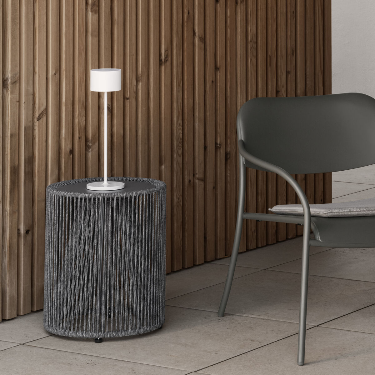 Eine mobile LED-Tischleuchte »FAROL« steht auf einem kleinen runden Beistelltisch neben einem grauen Sessel. Der Raum ist modern und minimalistisch eingerichtet mit Holzvertäfelung und großformatigen Fliesen.