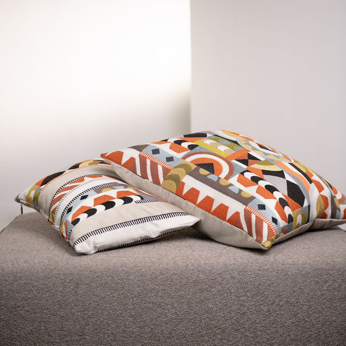 Zwei gemusterte Kissen im Havannah-Design liegen auf einem beigefarbenen Sofa. Die Kissen zeigen verschiedene geometrische Muster in Orange, Grün, Schwarz, Weiß und Beige.