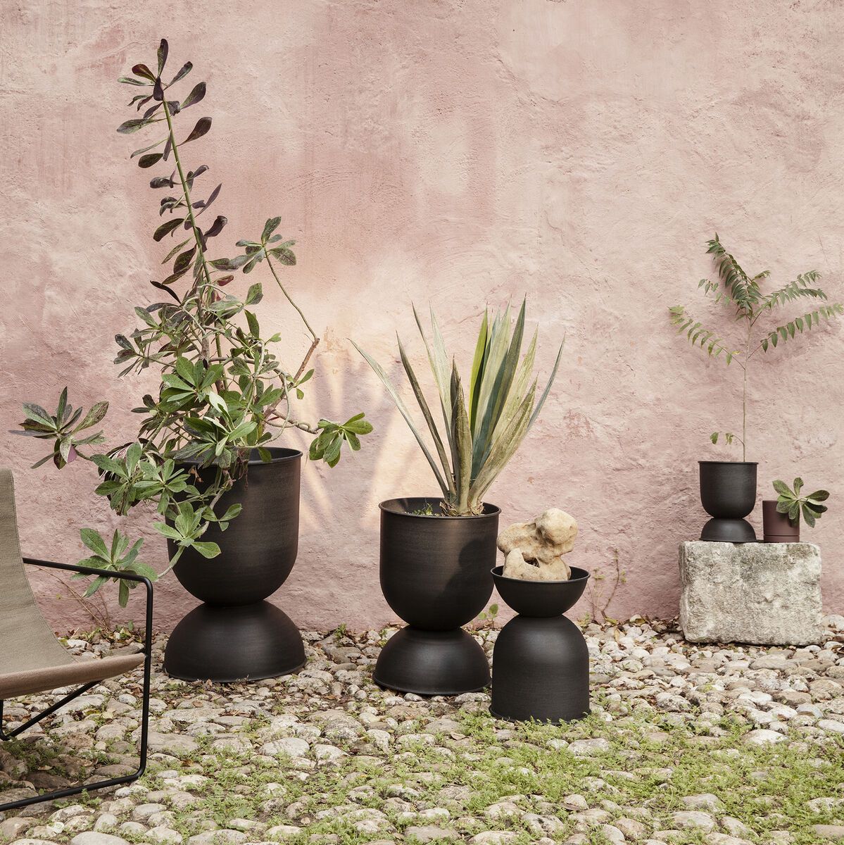 Mehrere dunkelgraue Pflanztöpfe der Serie »Hourglass« mit verschiedenen grünen Pflanzen stehen vor einer rosa Wand auf einem Kiesboden, daneben ein Metallstuhl.