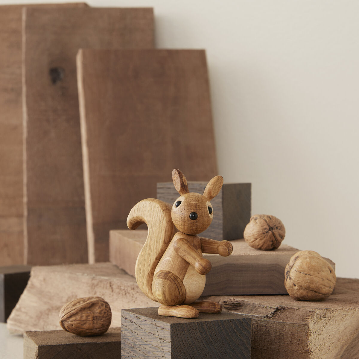 Handgefertigte Holzfigur eines Eichhörnchens namens 'Peanut', sitzend zwischen mehreren naturbelassenen Holzklötzen und Walnüssen; neutrale und warme Erdtöne dominieren das ruhige, minimalistische Arrangement.