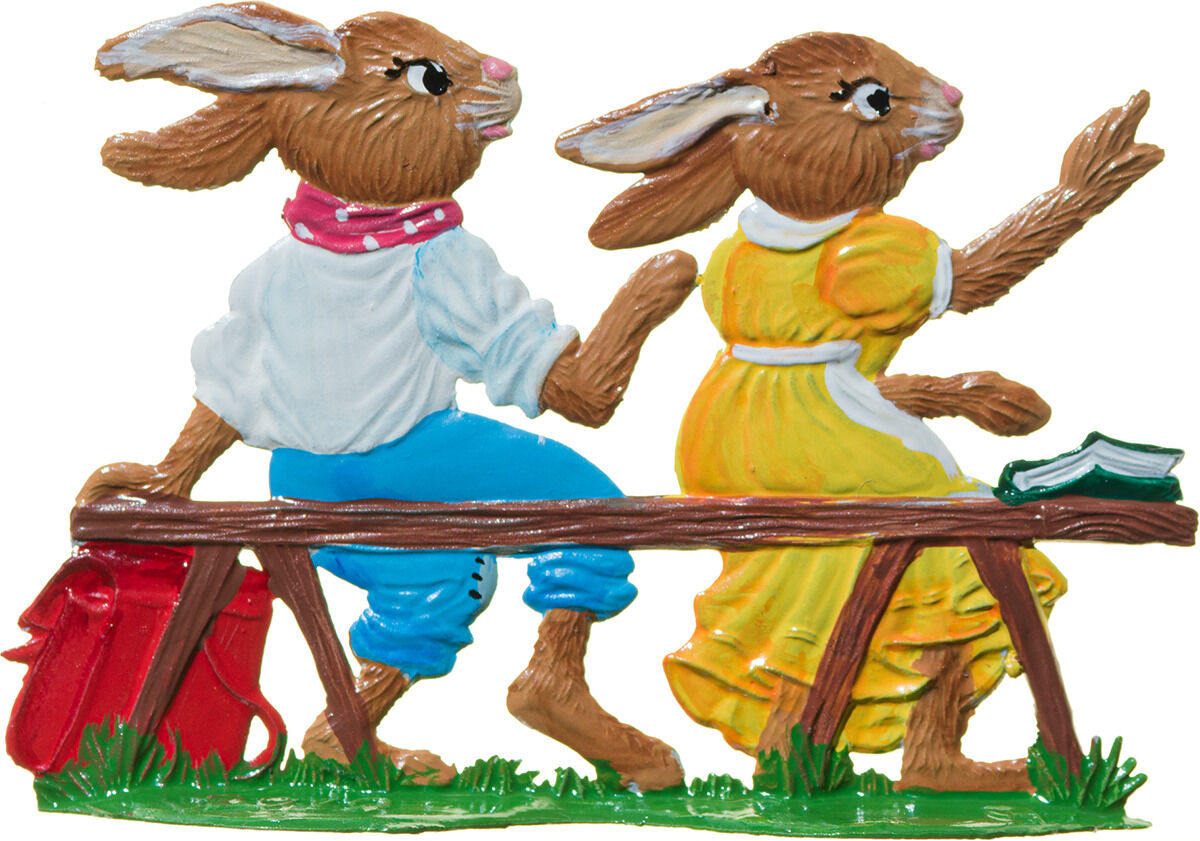 Zwei detailliert bemalte Zinnhasenfiguren der Marke Wilhelm Schweizer Zinnmanufaktur, ein Hase im weißen Hemd und blauer Hose, einer im gelben Kleid, gehen nebeneinander auf einer Wiese. Dabei trägt einer eine rote Tasche und ein grünes Buch liegt auf der Holzbank.