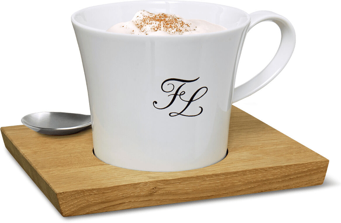Weiße Bürotasse mit Monogramm, präsentiert auf einem braunen Holzuntersetzer mit einem kleinen silbernen Löffel daneben. Die Tasse enthält eine weißliche Flüssigkeit mit einer braunen Pulverbestäubung oben drauf.