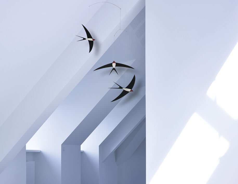Ein Mobile mit drei stilisierten Schwalben hängt an einer Decke. Die Schwalben sind schwarz mit weißen und roten Details. Der Hintergrund ist eine weiße, minimalistische Raumdecke mit schrägen Dachflächen.