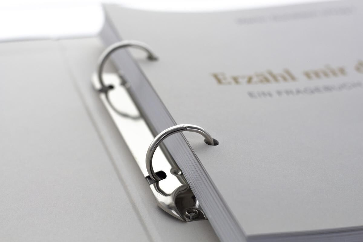 Ein gebundenes Erinnerungsbuch mit dem Titel 'Erzähl mir dein Leben' in der Leinenausgabe, geöffnet und mit einem Ringbuchmechanismus versehen. Die Seiten sind weiß, und der Titel ist in goldener Schrift gedruckt.