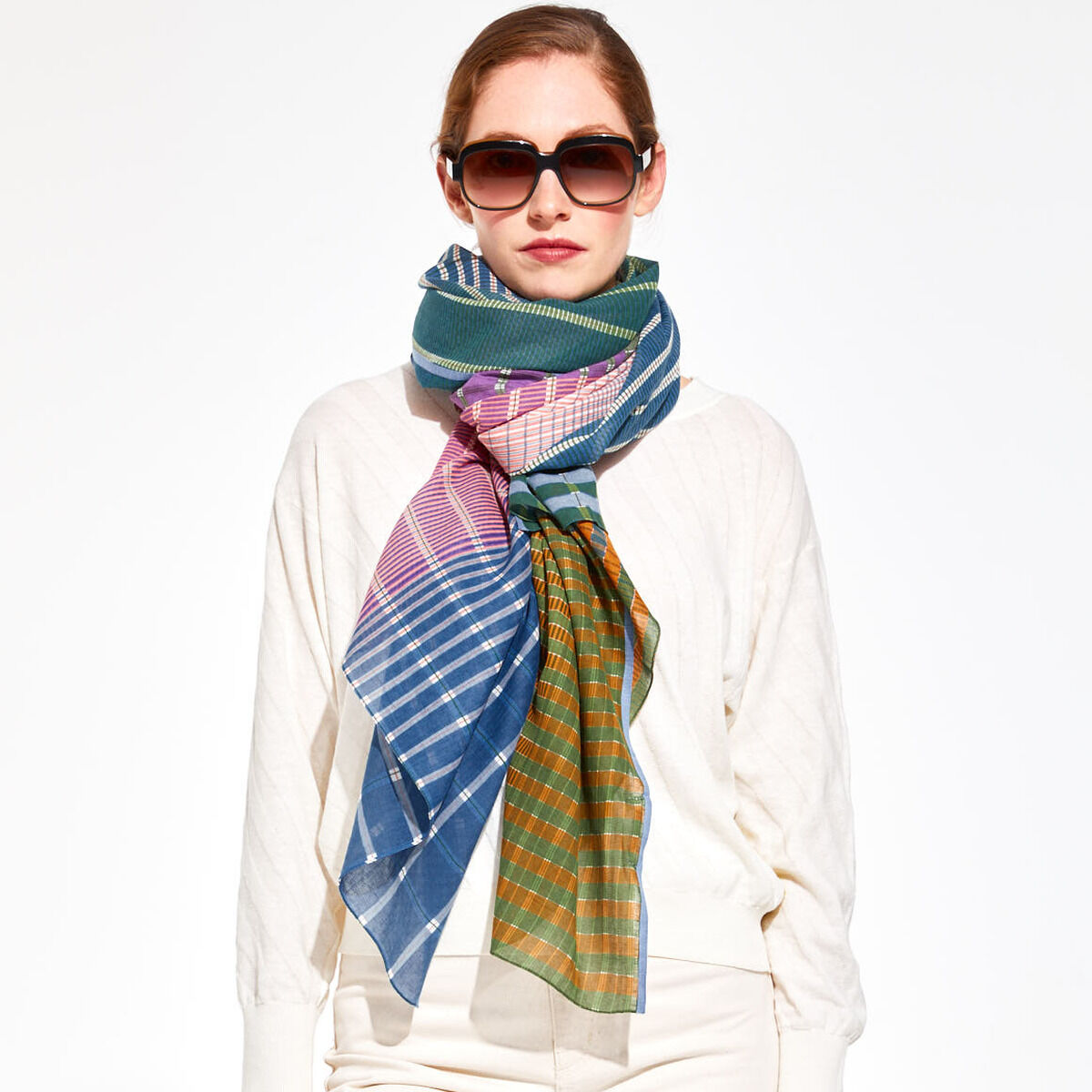 Frau trägt ein buntes Baumwolltuch mit Streifenmuster in Blau, Pink, Grün und Orange. Das Tuch ist um den Hals gewickelt und wird mit einer weißen Bluse kombiniert. Sie trägt Sonnenbrillen und posiert vor einem neutralen Hintergrund. Modisches Accessoire für Frühling oder Herbst.