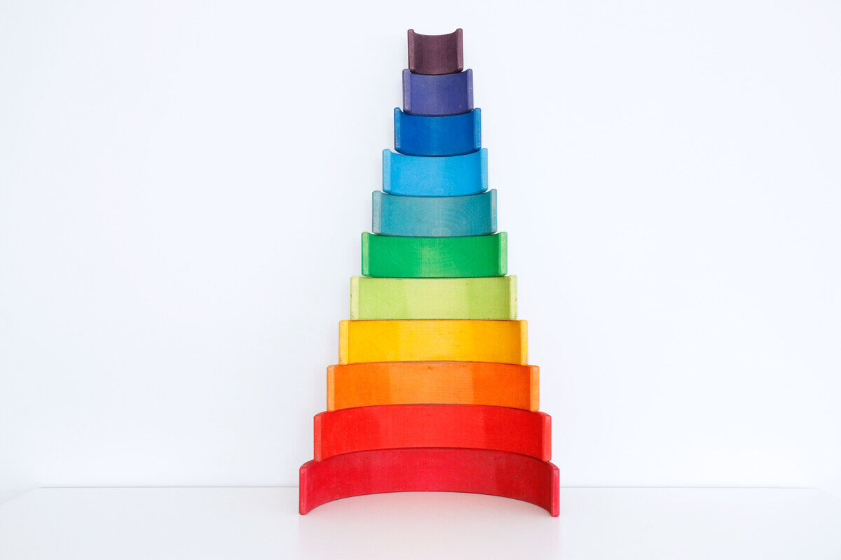 Ein Regenbogen-Holzspielzeug in verschiedenen Farben, geordnet in einem abgestuften Bogenmuster. Die Holzblöcke sind in den Farben Rot, Orange, Gelb, Grün, Türkis, Blau und Lila angeordnet.