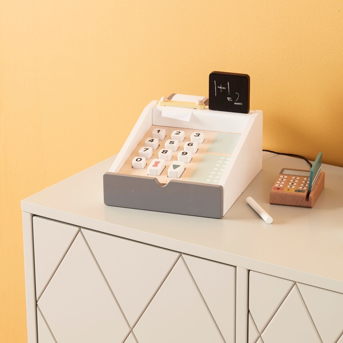 Spielzeugkasse aus Holz auf einem hellen, geometrisch gemusterten Schrank. Die Kasse hat einen grauen und weißen Körper mit schwarzen und weißen Tasten, einem kleinen Tafelbildschirm und einem Abakus aus Holz daneben.