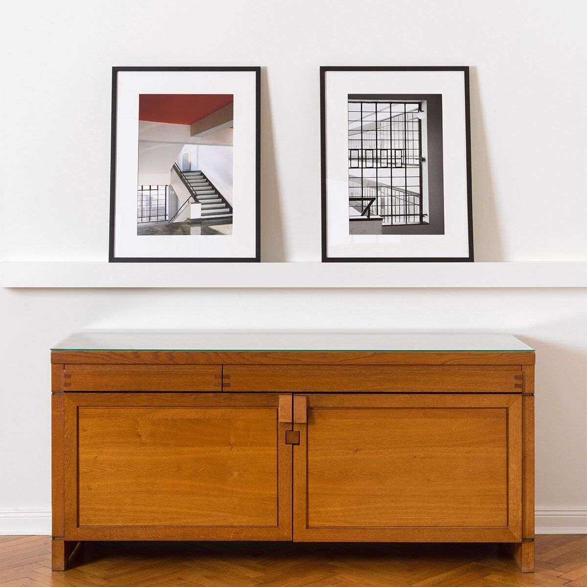 Zwei gerahmte Fotografien von Klaus Kinold, die das Haupttreppenhaus der Bauhaus-Schule in Dessau, entworfen von Walter Gropius, im Jahr 1992 zeigen. Die Fotografien stehen auf einem weißen Regal über einem Holztisch.