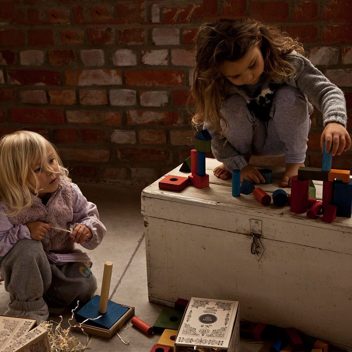 Zwei Kinder spielen mit Regenbogen Holzbausteinen in einem rustikalen Raum. Ein Kind baut auf einer weißen Holztruhe, während das andere am Boden spielt. Die Holzbausteine sind in verschiedenen Farben wie rot, blau, grün, gelb und orange.