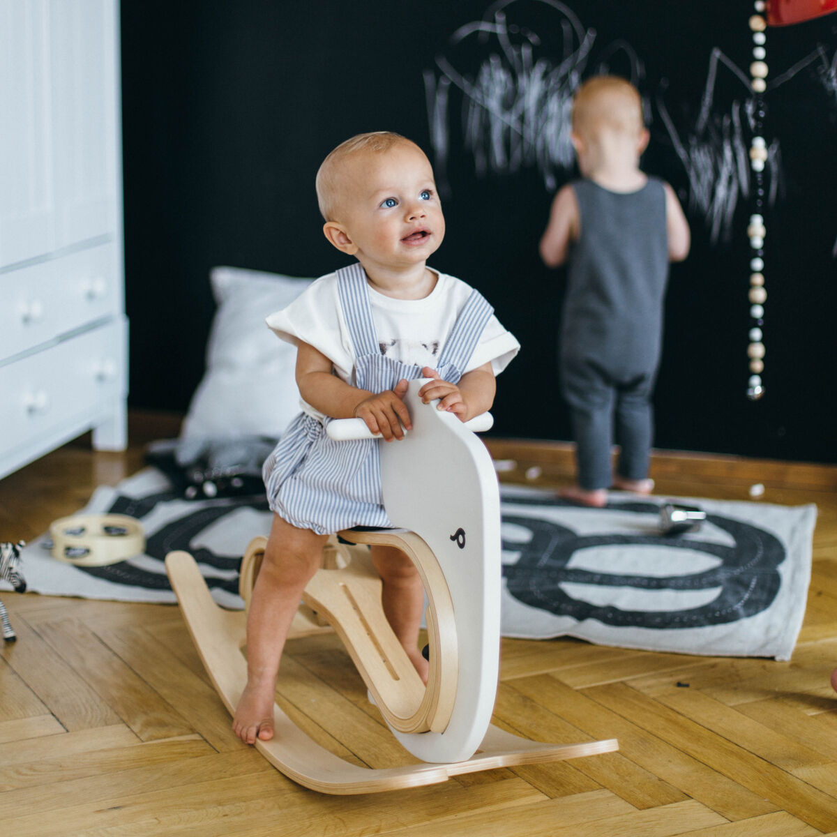 Ein kleines Kind sitzt auf einem weißen Schaukeltier in Elefantenform aus Holz in einem modern eingerichteten Kinderzimmer mit hellem Holzboden und schwarzer Wand. Ein weiteres Kind spielt im Hintergrund. Das Spielzeug wirkt hochwertig und ist für Kleinkinder geeignet.
