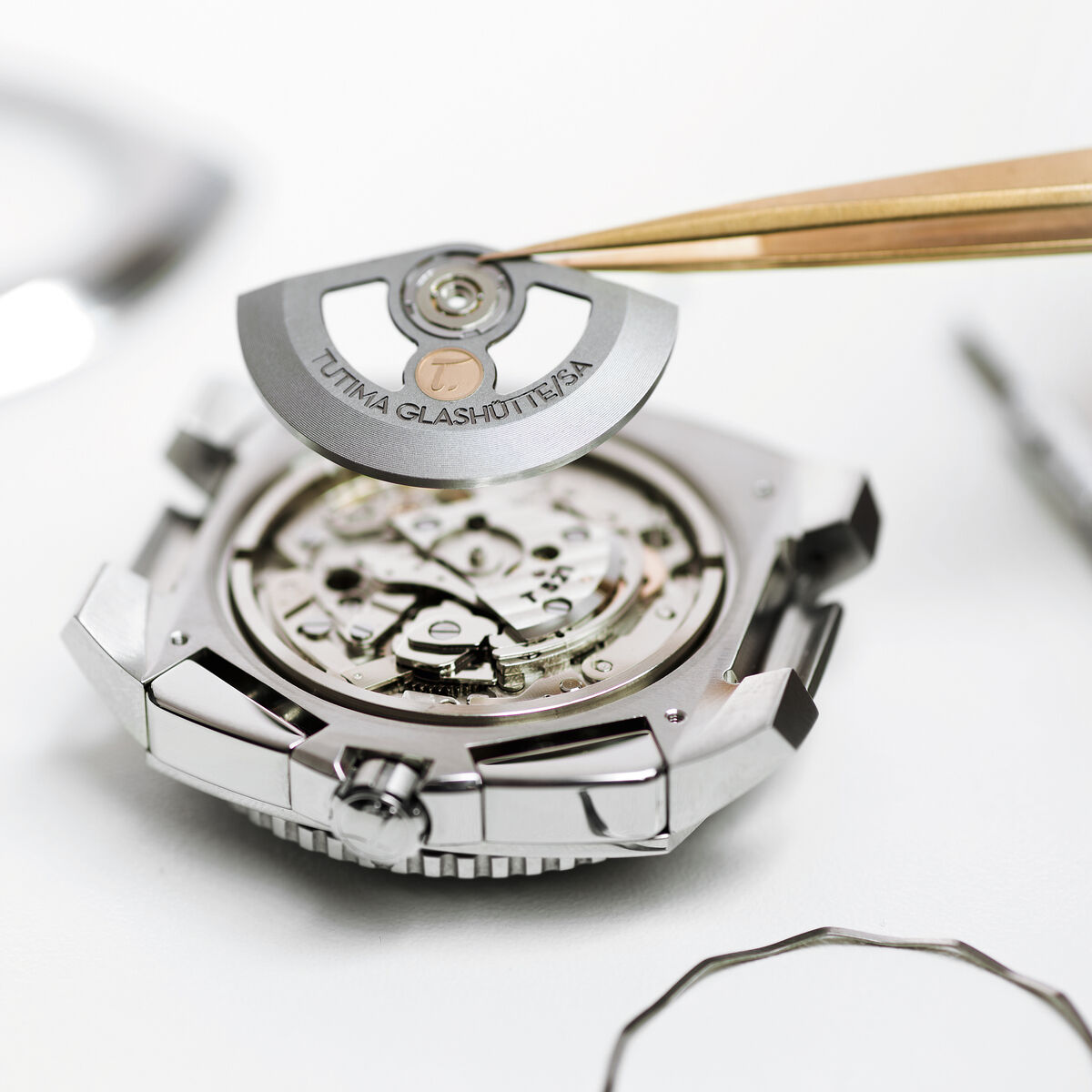 Eine hochwertige Tutima Glashütte Automatikuhr »Grand Flieger Airport Automatic« in der Montage: Das silberne Uhrwerk wird mit einer feinen Pinzette präzise eingesetzt. Im Hintergrund liegen Werkzeug, Uhrengehäuseteile und eine Werkhalterung auf weißer Unterlage.