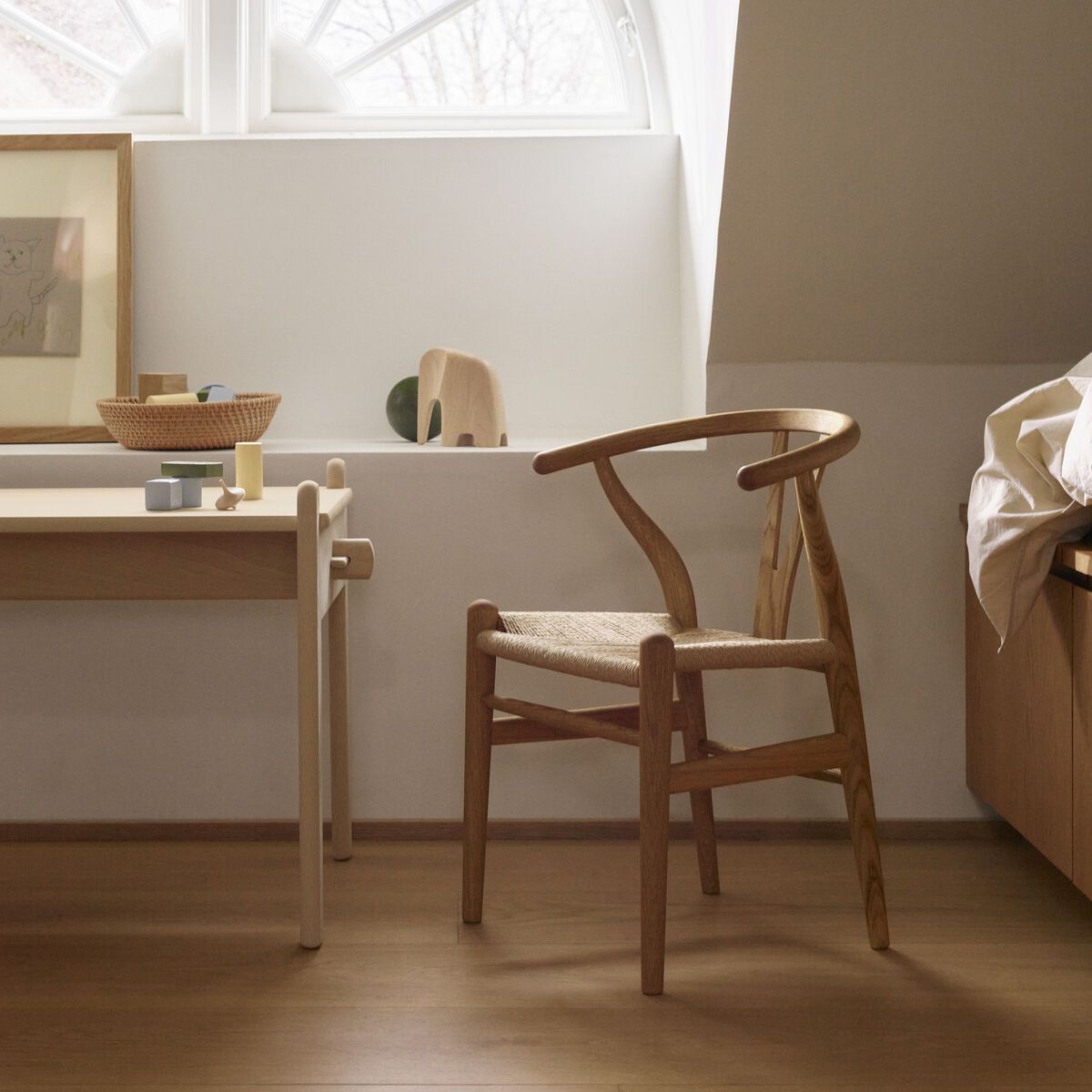 Ein stilvolles Kinderzimmer mit einem Holzstuhl »Wishbone Chair CH24 Kinder« und einem weißen Tisch unter einem runden Fenster. Der Raum ist mit naturholzfarbenen Möbeln und minimalistischer Dekoration eingerichtet.
