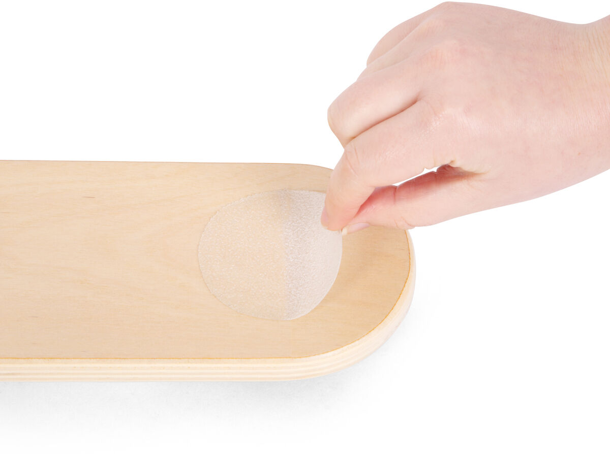 Eine Hand platziert einen runden, durchsichtigen Kunststoffaufkleber auf ein rechteckiges, hellbraunes Holzbrett.