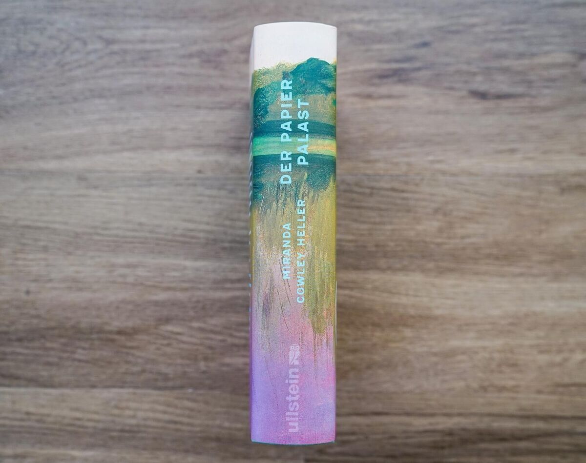 Eine gebundene Ausgabe des Romans 'Der Papierpalast' von Miranda Cowley Heller, verpackt in einer farbenfrohen Kunststoffhülle mit Grün-, Gelb- und Rosatönen, liegt auf einem Holztisch.