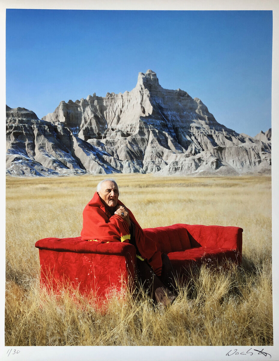 Ein älterer Mann sitzt in einem roten Sofa, das mitten in einer grasbewachsenen Prärie steht. Im Hintergrund erheben sich schroffe Berge unter einem klaren blauen Himmel.